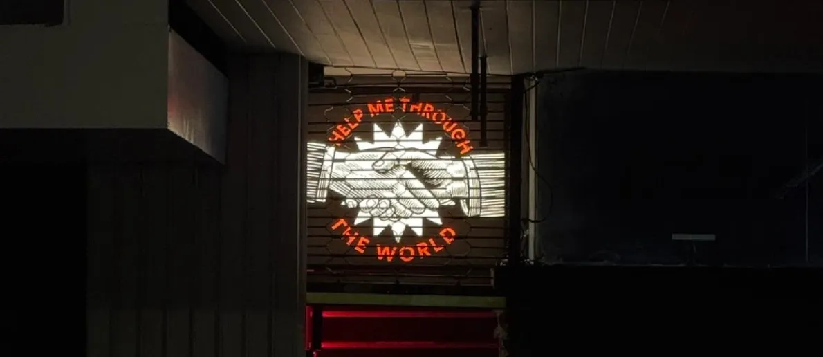 The illuminated Help Me Through the World sign, two clasped hands inside a starburst, glowing in a dark doorway