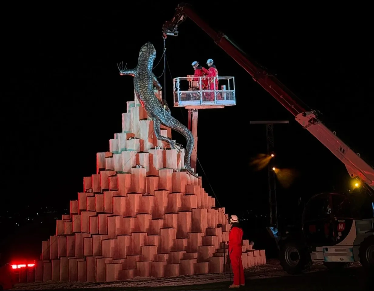 Workers in red overalls on a crane platform settle the skink onto a stepped pyre of cardboard boxes at night