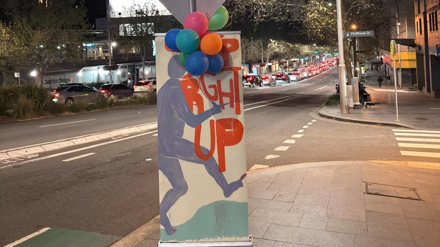 A hand-painted banner of a running figure, tied with balloons, on a city footpath at night with traffic streaming behind it