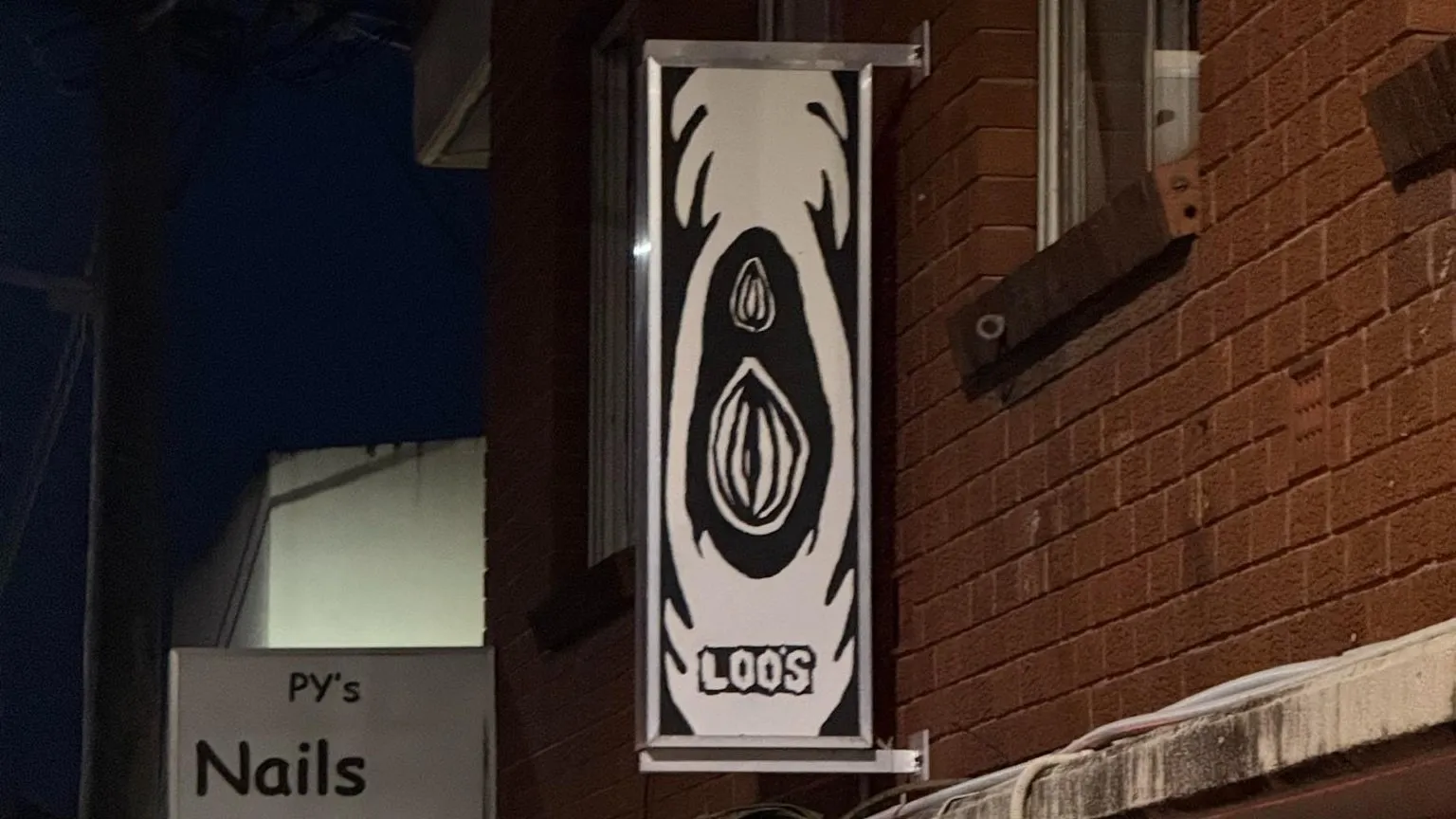 The Loo’s sign hanging from a brick terrace at night, above a nail salon sign